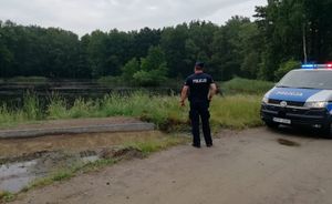 Policjant stoi przy zbiorniku wodnym i patrzy w jego stronę obok zaparkowany jest radiowóz.