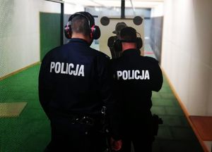 Dwóch policjantów stoi odwróconych tyłem, patrzą w stronę tarczy do strzelania, na uszach mają założone słuchawki.