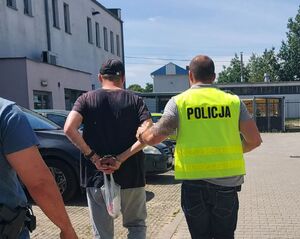 Policjant w żółtej kamizelce z napisem policja na plecach prowadzi zatrzymanego mężczyznę.