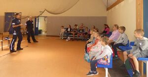 Policjantka i policjant stoją na środku sali a przed nimi siedzi grupa młodych ludzi.