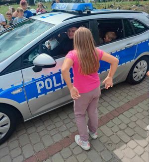 Dziewczynka stoi przy radiowozie za kierownicą którego siedzi policjant.