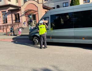 Policjant w żółtej kamizelce stoi przy szarym busie, zaparkowanym pod budyniem szkoły.