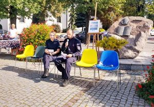 Policjantka i policjant siedzą na krzesełkach, policjant trzyma przy twarzy mikrofon.