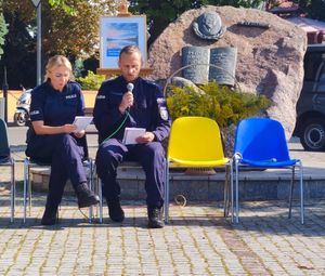 Policjantka i policjant siedzą na krzesełkach, policjant trzyma przy twarzy mikrofon.