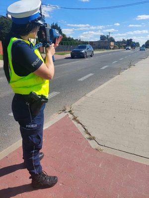 Policjantka w żółtej kamizelce stoi przy drodze i mierzy prędkość, ulicą przejeżdźają samochody.