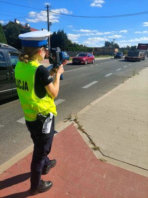 Policjantka w żółtej kamizelce stoi przy drodze i mierzy prędkość, ulicą przejeżdźają samochody.