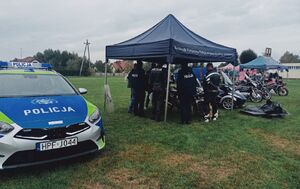 Policjanci stoją pod granatowym namiotem, obok stoi zaparkowany radiowóz i motocykl policyjny.