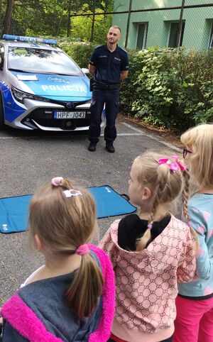 Policjant stoi przy radiowozie, przed nim stoją dzieci.