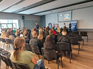 Policjant stoi na środku sali i mówi przez mikrofon do osób siedzących przed nim na krzesłach