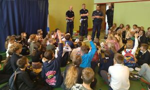 Trzech policjantów stoi na środku sali, przed nimi siedzi duża grupa dzieci.