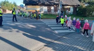 Policjanci stoją na drodze przez którą przechodzą dzieci w odblaskowych kamizelkach.
