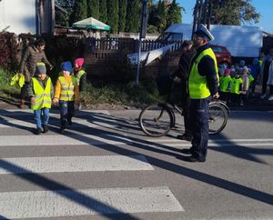 Policjanci stoją na drodze przez którą przechodzą dzieci w odblaskowych kamizelkach.
