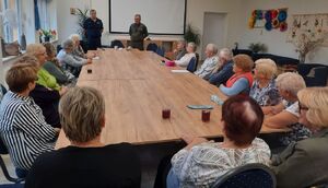 Policjant i strażnik leśny stoją na środku sali, w której przy owalnym stole siedzą seniorzy.