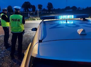 Dwóch policjantów stoi przy radiowozie, w tle widać cmentarz i palące się znicze.