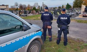 Dwóch policjantów stoi przy radiowozie zaparkowanym na cmentarzu.