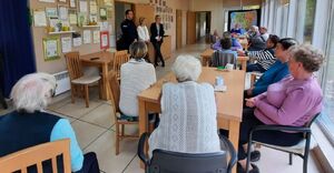 Policjant i dwie kobiety stoją na środku sali, w której przy stolikach siedzą starsze osoby.