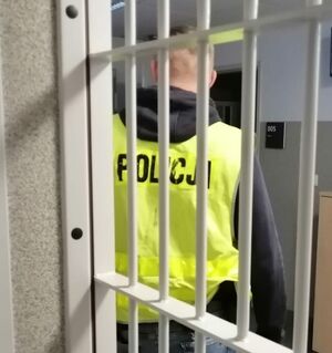 Policjant w żółtej kamizelce stoi za okratowanymi drzwiami.
