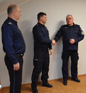 Trzech policjantów stoi obok siebie, jeden z nich podaje rękę policjantowi stojącemu w środku.