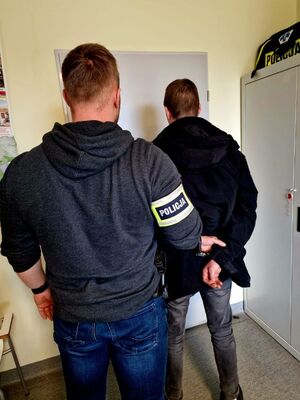 Policjant stoi obok zatrzymanego, który trzyma ręce zapięte z tyłu kajdankami.