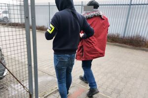Policjant prowadzi mężczyznę w czerwonej kurtce.