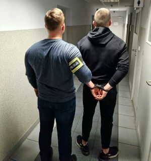 Policjant prowadzi zatrzymanego mężczyznę, który ma założone kajdanki na ręce trzymane z tyłu.