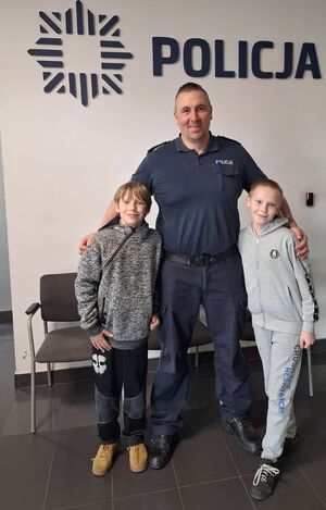 Policjant stoi pod napisem na ścianie Policja, obok niego stoi dwóch uśmiechniętych nastolatków.