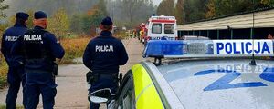 Policyjny radiowóz, obok niego stoi trzech policjantów, przed nimi stoi zaparkowana karetka pogotowia.