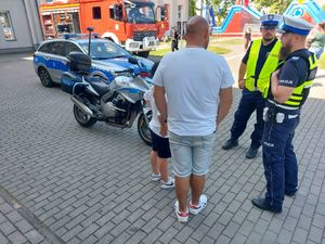 Dwóch policjantów stoi obok mężczyzny, za nimi stoi zaparkowany radiowóz, motocykl i wóz strażacki.