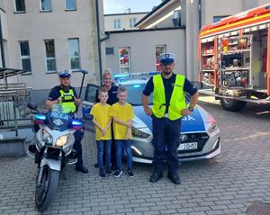 Dwóch policjantów i policjantka stoją obok dwóch chłopców w żółtych podkoszulkach, za nimi zaparkowany radiowóz i policyjny motocykl.