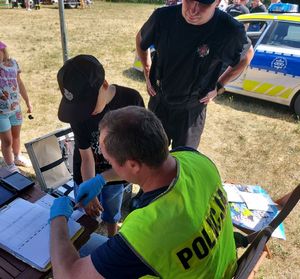 Policjant w żółtej kamizelce z napisem policja pobiera odciski palców od małego chłopca, obok nich stoi mężczyzna w czapce z daszkiem.