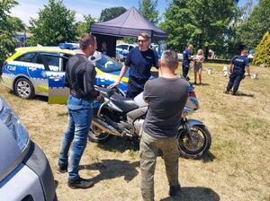 Policjanci stoją przy radiowozach i policyjnym namiocie i rozmawiają z mężczyznami i kobietami.