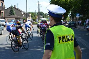 Policjant w białej czapce i żółtej kamizelce z napisem policja patrzy w stronę jadących kolarzy.