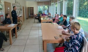 Policjant stoi na środku sali, obok niego siedzi policjantka a przed nimi siedzi grupa seniorów.