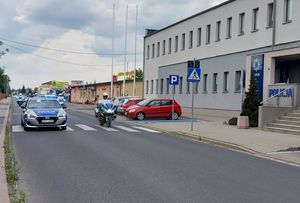 Policyjne motocykle i radiowozy jadą jezdnią obok budynku z napisem Policja.