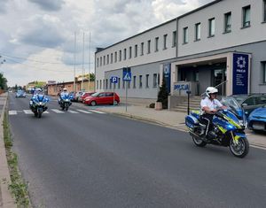 Policyjne motocykle i radiowozy jadą jezdnią obok budynku z napisem Policja.