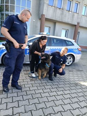 Dwóch policjantów stoi obok kobiety trzymającej psa, za nimi stoi zaparkowany radiowóz.