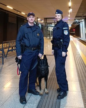 Policjantka i funkcjonariusz kolei trzyma psa, oboje chodzą po dworcu kolejowym.