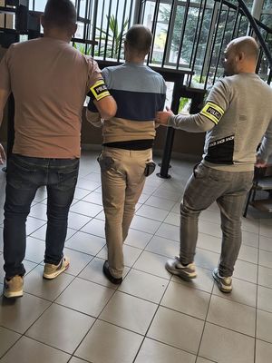 Dwóch policjantów z opaskami na przedramieniu z napisem policja prowadzą w środku zatrzymanego mężczyzny.