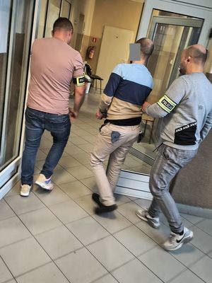 Dwóch policjantów z opaskami na przedramieniu z napisem policja prowadzą w środku zatrzymanego mężczyzny.