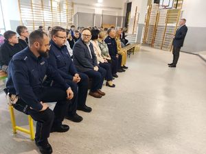 Policjanci siedzą na ławkach w sali sportowej z grupą innych osób.
