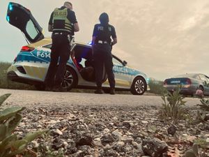 Policjanci stoją przy radiowozie, w którym siedzi zatrzymany mężczyzna.