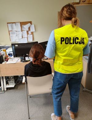Policjantka stoi za zatrzymaną kobietą, która siedzi przed policjantką na krześle.