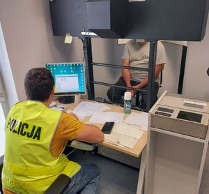 Policjant siedzi przed komputerem, przed nim siedzi zatrzymany mężczyzna.