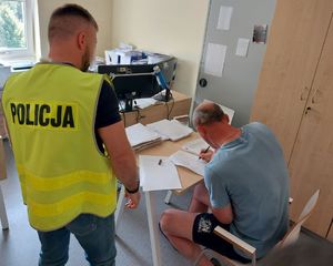 Policjant stoi przy zatrzymanym mężczyźnie, który siedzi przy stoliku i podpisuje dokumenty.