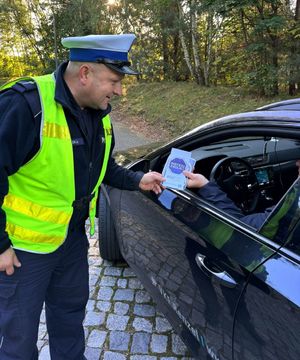 Policjant wręcza ulotki z napisem Mistrzu zwolnij kierującym autami.
