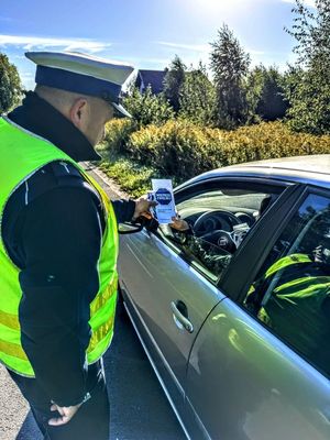 Policjant wręcza ulotki z napisem Mistrzu zwolnij kierującym autami.