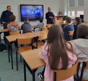 Dwójka policjantów stoi na środku klasy, przed nimi siedzą uczniowie w ławkach.