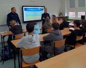 Dwójka policjantów stoi na środku klasy, przed nimi siedzą uczniowie w ławkach.