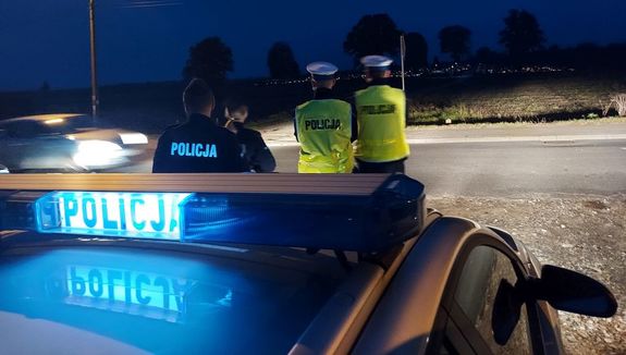 Policjanci stoją przed radiowozem, na wprost nich widać cmentarz.