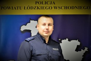 Policjant w granatowym mundurze stoi na tle kolorowego baneru.